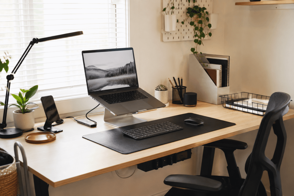 Small home office desk setup with a laptop stand, wireless mouse, desk lamp, cable management accessories, and a clean organized workspace in natural daylight.